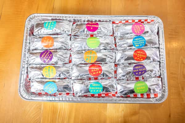 Overhead view of foil-wrapped tacos with colorful labels arranged in a rectangular catering tray.