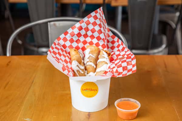 Chicken flautas drizzled with white sauce in a paper-lined cup, with orange dipping sauce alongside.