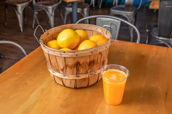 Basket of whole oranges and a clear cup of orange juice on a wooden restaurant table.