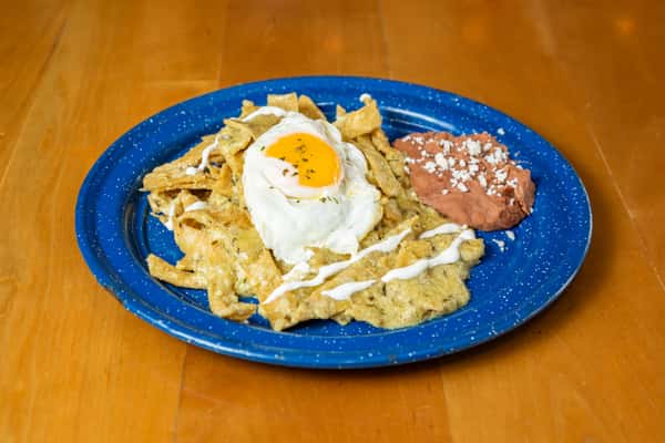 Chilaquiles topped with a fried egg and white sauce, with refried beans on a blue plate.