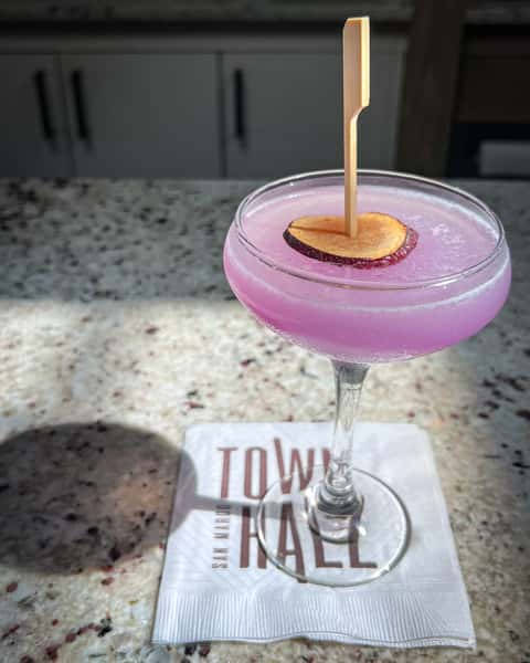 Purple cocktail in a coup glass at Town Hall with a skewer through the garnish. Town Hall cocktail napkin under glass, on bar.