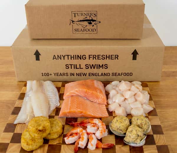raw, fresh haddock, salmon, scallops & stuffed clams, fish cakes and cocktail shrimp, all on a wooden board with Dock to Door shipping boxes behind.