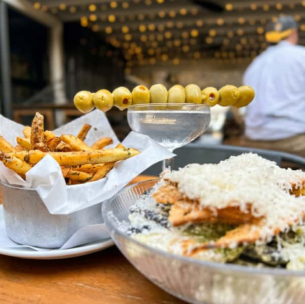A caesar salad topped with a layer of grated cheese and creamy dressing, served alongside a basket of golden fries. A chilled martini garnished with olives adds a refined touch, blending comfort food with an elevated dining experience.