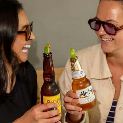 Two women clinking Mexican beer bottles garnished with fresh lime wedges—Modelo and a dark cerveza—smiling in a lively taqueria setting, capturing a fun happy hour moment with authentic Mexican drinks.