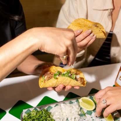 Handheld corn tortilla taco filled with seasoned meat, topped with fresh chopped cilantro and white onion, served with lime wedges—authentic street-style tacos in a lively Mexican restaurant.
