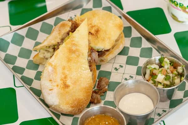 Toasted Mexican torta roll stuffed with seasoned carne asada, served with pico de gallo, creamy salsa, and house-made hot sauce on a metal tray—hearty street-style sandwich in a vibrant taqueria setting.