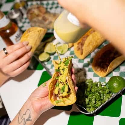 Handheld corn tortilla taco filled with seasoned carne asada, topped with fresh cilantro and drizzled with creamy green salsa, served with chopped onion and lime wedges—authentic Mexican street taco in a vibrant taqueria setting.