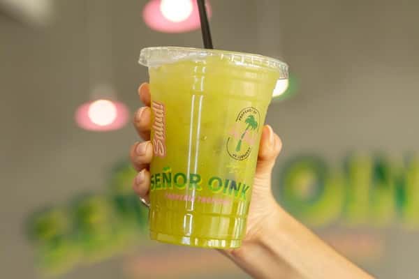 Iced green agua fresca served in branded cup with straw—refreshing Mexican fruit drink in a colorful, modern taqueria ambiance.