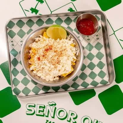Cup of Mexican street corn topped with crumbled queso fresco, chili powder, fresh lime wedge, and side of red salsa—classic elote flavors in a casual, colorful taqueria atmosphere.