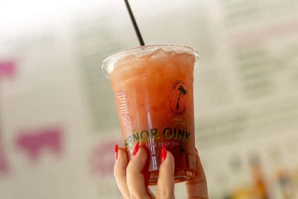 Iced watermelon agua fresca served in a Señor Oink branded cup, held up against a colorful taqueria backdrop—refreshing Mexican fruit drink with vibrant tropical flair.