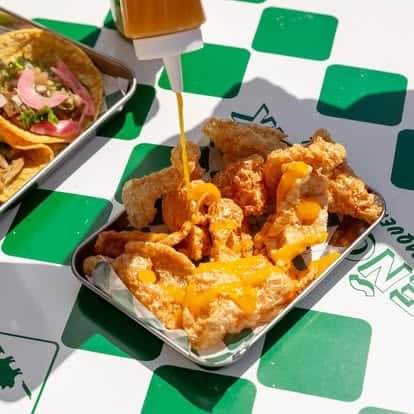Golden, house-made chicharrones topped with warm nacho cheese sauce, served on a metal tray with green checkered liner—bold Mexican street food flavors in a vibrant taqueria setting.