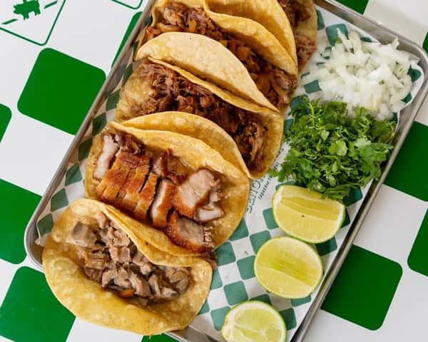 Tray of authentic street tacos filled with carnitas, crispy pork belly, and seasoned meats, topped with chopped onion and cilantro, served with fresh lime wedges.