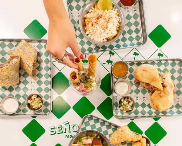 Overhead view of a Mexican taqueria spread featuring crispy rolled taquitos dipped in guacamole and pickled onions, a stuffed quesadilla, a torta sandwich with slow-cooked pork, elote topped with crumbled queso fresco and chili powder, plus fresh salsas and pico de gallo on green checkered trays in a vibrant fast-casual restaurant setting.
