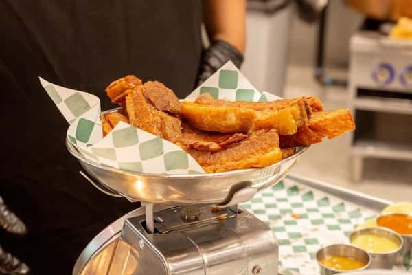 Close-up of crispy chicharrón strips and slow-roasted pork belly stacked in a metal bowl lined with green checkered paper, displayed on a countertop scale in a vibrant Mexican taqueria kitchen with house-made salsas in the background.