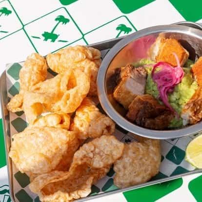 Crispy pork belly bites over creamy guacamole, garnished with pickled red onions and fresh cilantro, served alongside airy chicharrones—authentic Mexican appetizer with rich, savory flavor.