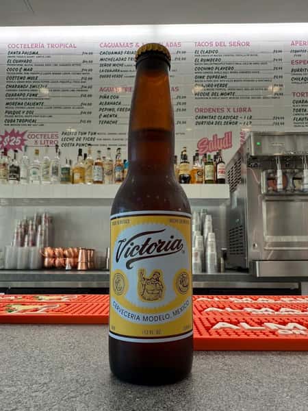 Victoria cerveza bottle displayed on bar counter with full cocktail menu backdrop—traditional Mexican beer complementing bold street food flavors.