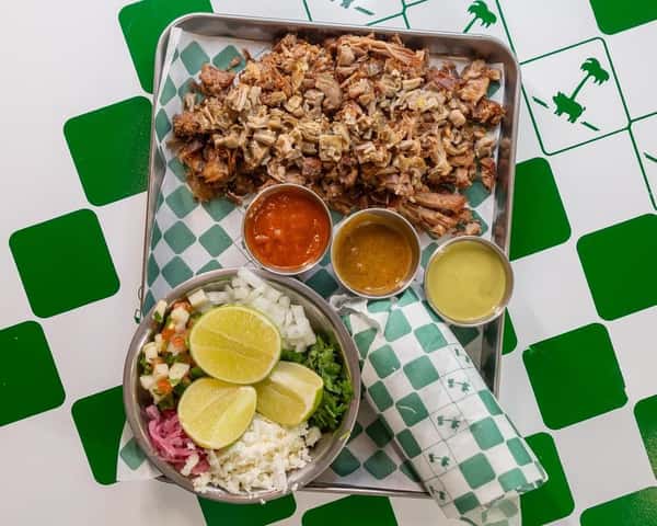 Tray of chopped carnitas served with fresh lime wedges, cilantro, onion, pickled red onions, queso fresco, and trio of house-made salsas—authentic Mexican feast perfect for sharing.