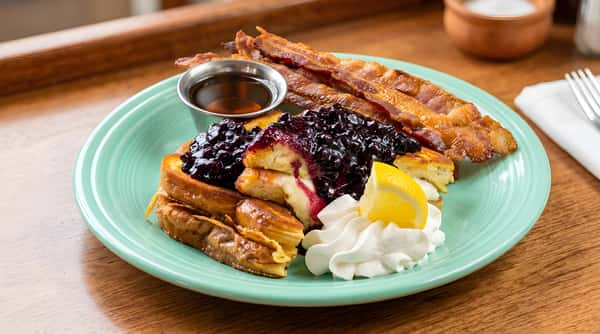 French toast topped with bacon, syrup, and whipped cream on a green plate.