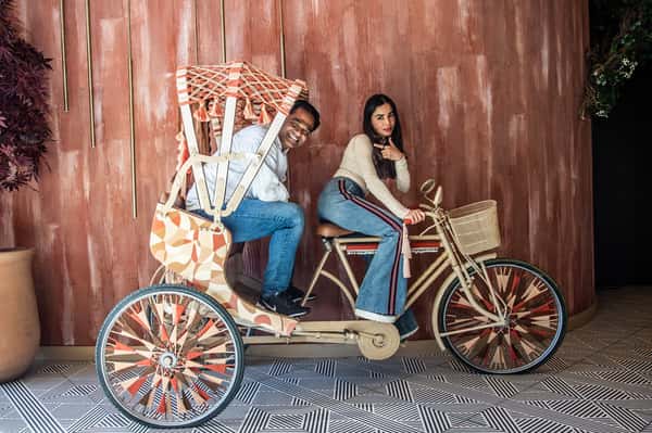 Two guests posing on a decorative vintage-style bicycle display inside an upscale tropical-inspired restaurant with playful modern ambiance