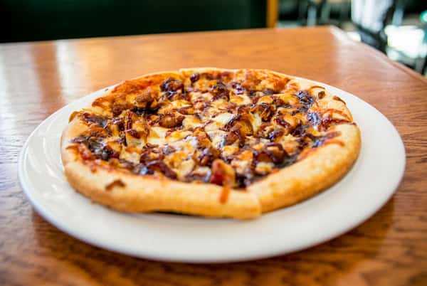 BBQ chicken pizza on a white plate, with melted cheese, chicken pieces, and BBQ sauce drizzle on a wooden table.