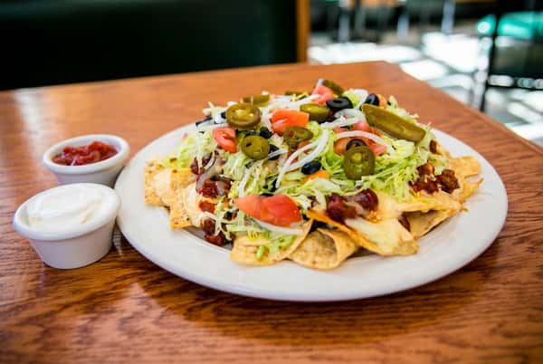 Loaded nachos with ground beef, cheese, shredded lettuce, tomatoes, onions, olives, and jalapeños. Salsa and sour cream are in small bowls.