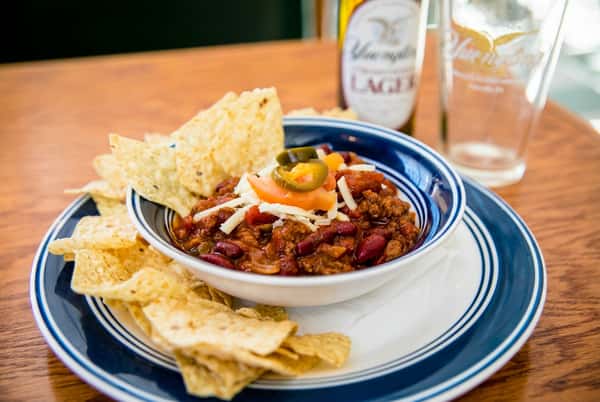 Bowl of chili with shredded cheese, sliced tomatoes, and jalapeños, surrounded by tortilla chips.