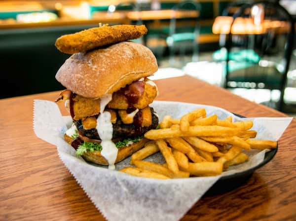 A large hamburger loaded with bacon, fried onion rings, lettuce, tomato, and sauce, served with french fries.