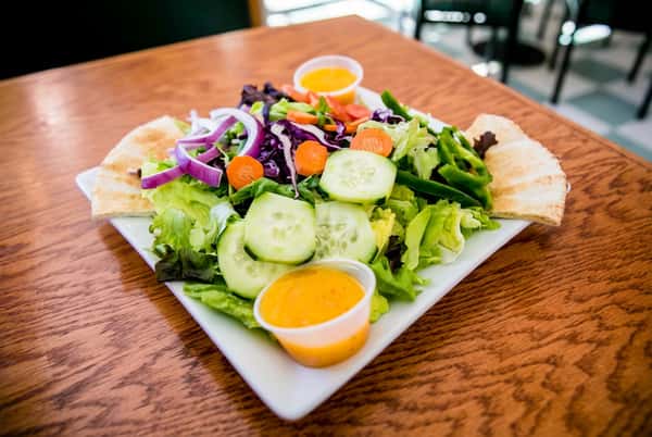 Garden salad with pita bread, mixed greens, red onion, carrots, cucumbers, and two cups of orange dressing on a white plate.