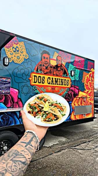 Hand holding a plate of tacos in front of the Dos Caminos Food Truck