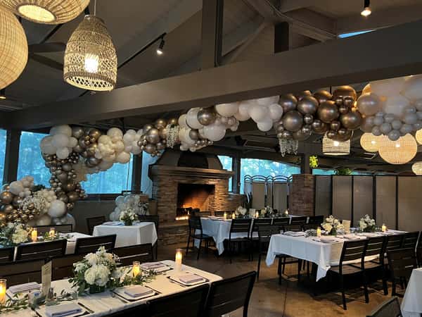 Evening event setup in a restaurant with romantic lighting, white linens, and lush white floral arrangements, highlighted by an overhead garland of metallic and pearl balloon clusters for a glamorous touch.