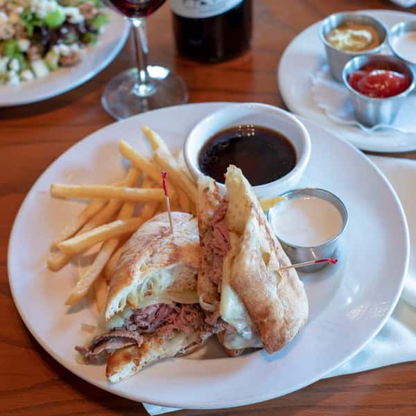 A plate of golden crispy French fries served alongside a toasted club-style sandwich filled with layers of deli meat, lettuce, and melted cheese. Several dipping sauces and a glass of red wine complement the dish, making it a satisfying casual meal.