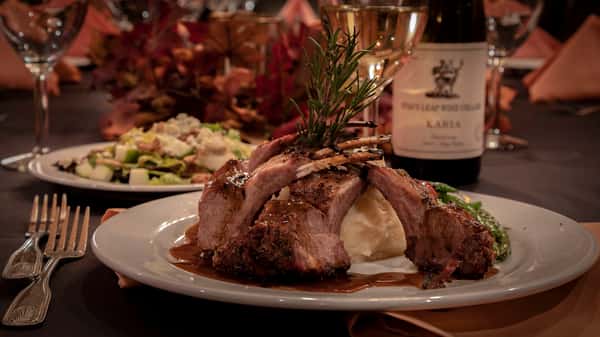 A beautifully plated rack of lamb cooked to perfection, served over creamy mashed potatoes and garnished with a fresh sprig of rosemary. The dish is surrounded by roasted vegetables and paired with a glass of white wine, creating an elegant and upscale dining presentation.