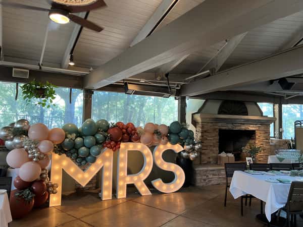 Rustic restaurant corner with large marquee "MRS" letters framed by pastel and metallic balloons, perfect for a bridal shower or wedding celebration, set beside a stone fireplace and large windows.