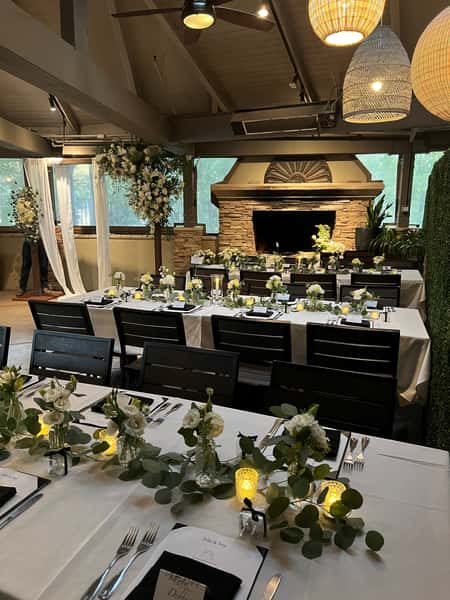 Sophisticated wedding reception setup in a rustic restaurant venue, with white linens, soft greenery, glowing votive candles, and white floral arrangements in front of a stone fireplace and floral arch.