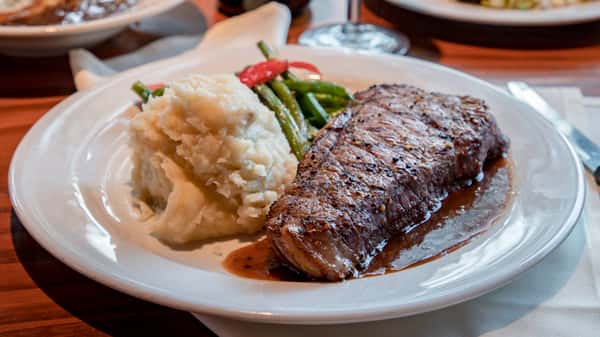 A juicy grilled steak cooked to perfection and served with creamy mashed potatoes and a side of fresh green beans. The steak is topped with a rich savory sauce, creating a classic and hearty steakhouse-style meal.