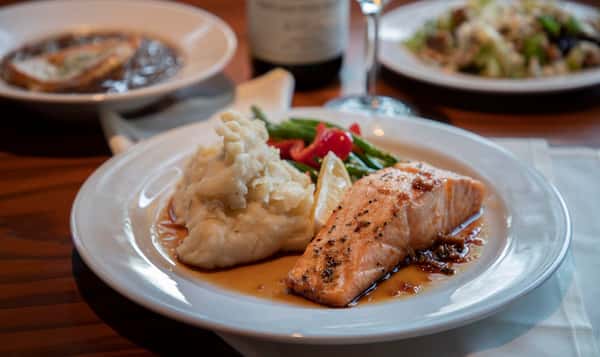 A perfectly seared salmon fillet served with creamy mashed potatoes and a side of crisp green beans and roasted red peppers. The dish is drizzled with a savory sauce, offering a comforting and flavorful restaurant-style entrée.