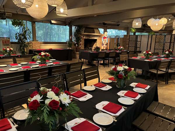 Elegant indoor restaurant space set for a holiday event, featuring black tablecloths, red napkins, and festive floral centerpieces with red and white roses, surrounded by wooden chairs and warm lighting.
