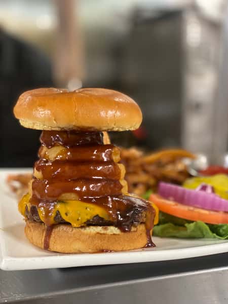 A gourmet burger stacked high with crispy onion rings, melted cheese, and barbecue sauce on a glossy brioche bun, served with fresh garnishes in the background.