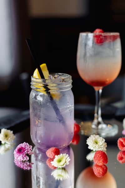 A colorful cocktail pairing featuring a sparkling lavender drink in a mason jar and a fruity citrus cocktail garnished with fresh berries and an orange twist.
