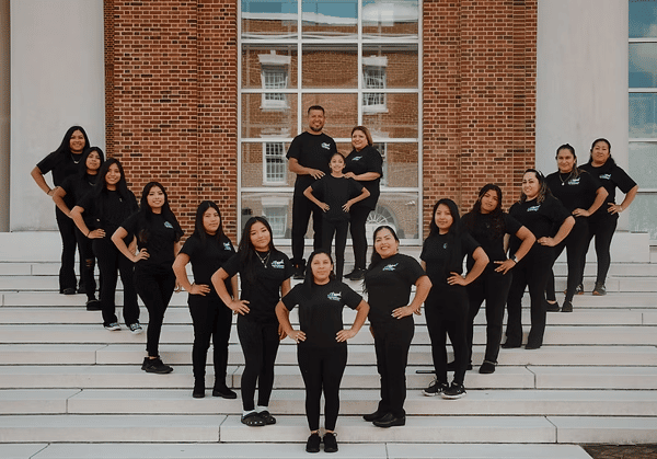 A group of restaurant staff in matching black uniforms posing together on white steps in front of a brick building with large windows.