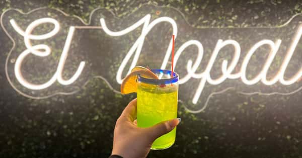 A hand holding a bright green cocktail garnished with an orange slice in front of an illuminated "El Nopal" sign.