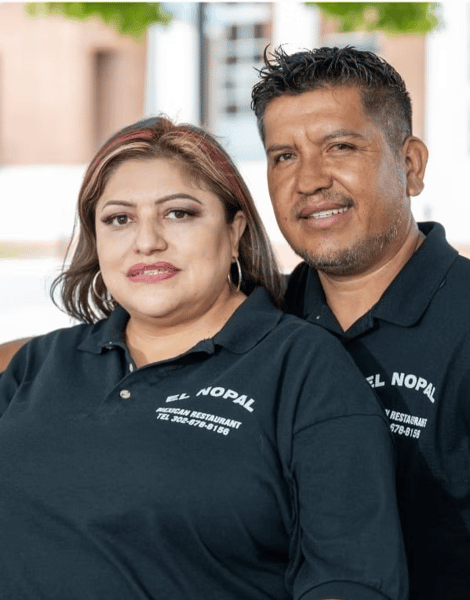 Two people wearing matching "El Nopal Mexican Restaurant" polo shirts, smiling and posing closely together outdoors.