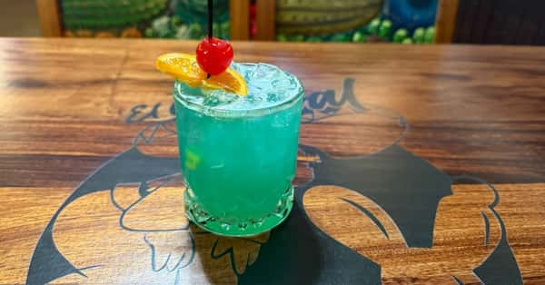 A vibrant blue cocktail served over ice in a short glass, garnished with a cherry and an orange slice on a skewer. The drink is presented on a wooden table featuring the "El Nopal" logo.
