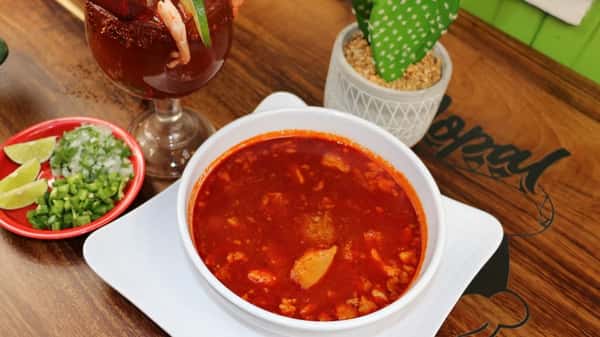 Bowl of red pozole served with lime, chopped onion, cilantro, jalapeños, and a shrimp cocktail in a michelada glass.