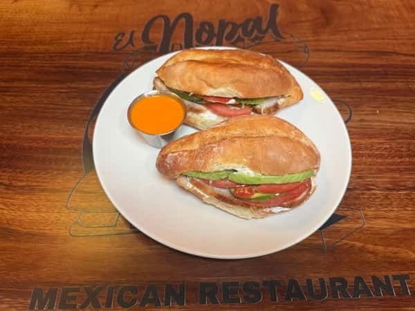 Two Mexican tortas filled with sliced avocado, tomato, and what appears to be a creamy spread or cheese, served with a side of orange-colored dipping sauce.