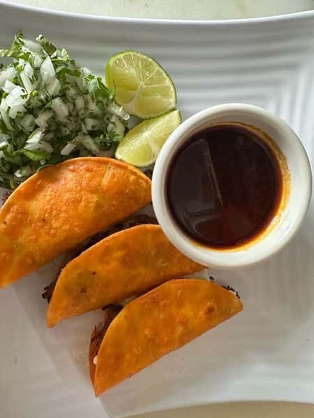 Birria tacos with a side of chopped onions, cilantro, lime wedges, and a cup of rich consommé for dipping.