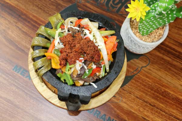 Molcajete dish filled with grilled beef, chorizo, sautéed bell peppers, onions, and melted cheese, garnished with cactus paddles.