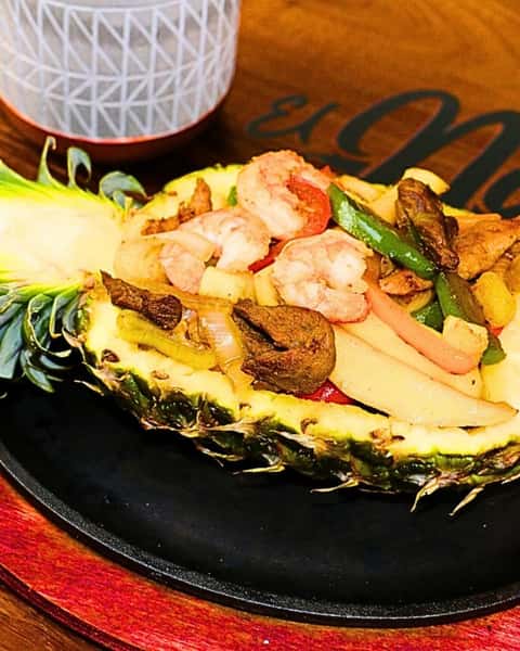 This dish is a **pineapple fajita bowl**, served in a halved pineapple. It contains a vibrant mix of grilled shrimp, beef, bell peppers (green and red), onions, and pineapple chunks. The pineapple base adds a tropical sweetness that complements the savory and slightly spicy fajita-style ingredients.