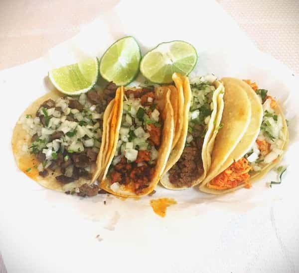 Five street tacos with assorted meats, topped with onions and cilantro, served with lime wedges on a white tray.