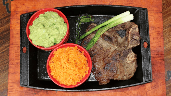 Grilled T-bone steak served with guacamole, Mexican rice, and grilled green onions on a cast iron plate.
